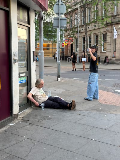 The image shows a street scene in Nottingham, United Kingdom. An elderly man is lying on the pavement next to a shop front with a "Fire Exit, Keep Clear" sign. He is wearing a light-colored t-shirt and dark trousers. A plastic water bottle and a cup are near him on the ground. A person wearing a black t-shirt and very baggy blue jeans is standing on the pavement drinking from a bottle of water. Two other people are walking on the pavement. A traffic light pole is positioned near the shop front. Buildings are visible in the background, including one with a "RED DOG" sign.
