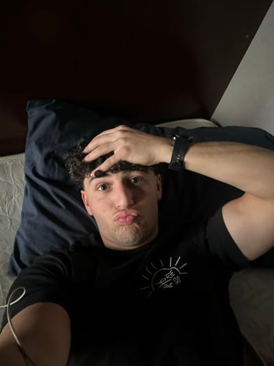 A young adult male is depicted in a close-up, upward-angled self-portrait, likely taken in a bedroom within the city of Sarouja Municipality, Syria. The individual has dark, curly hair and brown eyes, and is looking directly at the camera with his lips pursed. His right hand rests on his head, fingers touching his hair. He is wearing a black short-sleeved t-shirt with a white graphic emblem on the chest, featuring a stylized sun and "NIKE SB" text. A black digital wristwatch is present on his left wrist. He lies on his back, his head resting on a dark blue pillow, which in turn lies on a white, textured mattress or bedspread. The background consists of a dark brown surface, possibly a headboard or wall, suggesting an indoor, low-light environment. A white cable is visible across the lower left corner of the frame.