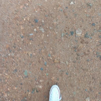 The image depicts a ground surface composed of reddish-brown dirt and small pebbles. A portion of a white tennis shoe with a red stripe is visible in the lower right corner of the frame. The image appears to be taken from a first-person perspective, looking down at the ground. There are small rocks in the dirt. It can not be determined that the location is in Mahiakalo ward, Kenya, as there is no evidence of that location in the image.