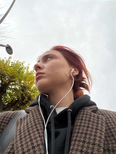 An outdoor, low-angle portrait depicts a young female subject positioned to the right of the frame, looking upwards and slightly to her right. She has reddish-brown hair and pale skin, with light pink lipstick. White wired earbuds are visible, with one inserted into her right ear, which also features a small silver stud earring. She is dressed in a black hooded sweatshirt beneath a brown and black patterned coat. A gray strap from a bag is visible over her left shoulder. In the background, an overcast gray sky dominates the upper portion of the image. To the left, a street light fixture is visible on a pole, and a tree with green and yellow autumnal leaves is present behind the subject. The scene is located in an urban environment within the city of Reims, France.