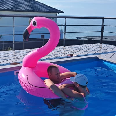 In a bright, sunlit outdoor swimming pool, a man and a woman are enjoying a relaxing day. They are both partially submerged in the clear blue water, leaning against a large, inflatable pink flamingo pool float. The man, with short, light-colored hair, looks towards the side, while the woman, wearing a white baseball cap and a pink bikini, leans her head on his shoulder. The flamingo float is the central object, its neck arched and head held high, with a cartoonish eye and beak. The pool is surrounded by a wooden deck with a dark railing. In the background, a modern building with a dark tiled roof and large windows is visible, suggesting a residential or hotel setting. Beyond the building, the calm blue sea stretches to the horizon under a clear sky, indicating a coastal location. The overall atmosphere is one of leisure and happiness. The time of day appears to be midday, given the bright sunlight and shadows.