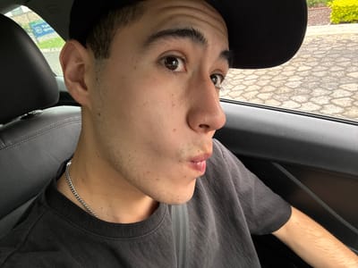 The image captures a young male situated in the passenger seat of a vehicle, in what is identified as San Bernardino Tlaxcalancingo, Mexico. The individual, with light to medium complexion, dark hair, and visible facial stubble, wears a dark t-shirt and a silver-toned cuban link chain. A dark baseball cap is partially visible on his head. His dark brown eyes are wide and directed towards the viewer, while his lips are pursed into a distinct pouting or kissing expression. A seatbelt is diagonally secured across his chest. The car's interior features dark grey leather seating and dark grey plastic door trim. Through the vehicle's windows, exterior elements are visible: on the left, behind a dark headrest, a distant, partially obscured green and blue sign, possibly indicating "Park" or "Parking," is discernible amidst green foliage and a damp-appearing road surface. On the right, a textured ground of interlocking light-colored pavers or cobblestones is brightly lit, contrasting with a vibrant yellow-green bush beyond it.