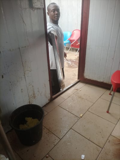 A man stands in a doorway, holding a flat, silver object. Behind him, an outdoor area with red and blue chairs is visible. The interior space has tiled flooring and walls of what appear to be prefabricated metal. A black bucket containing yellow objects sits in the corner. A red plastic chair is positioned at the edge of the frame. The room is somewhat cluttered with small pieces of debris scattered on the floor.
