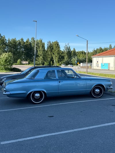 A light blue vintage four-door sedan is parked in an asphalt parking lot under a clear blue sky. The car has white-walled tires and chrome hubcaps. In the background, there is a line of green trees, a modern building with a red roof, and a utility pole with a street lamp. To the left of the vintage car, a dark-colored modern sedan is parked. The scene is likely in the city of Imatra, Finland.