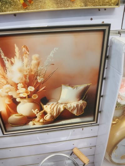 This media file showcases a framed artwork with a floral and still life theme. The artwork depicts a collection of dried grasses and flowers in a vase, placed alongside a woven basket containing a pillow and draped fabric. There are also several small ceramic or clay objects within the basket, resembling miniature jugs or pots. The color palette is predominantly warm, with shades of orange, brown, and cream, creating a soft and inviting atmosphere. The edges of the artwork and the decorative elements within the basket are outlined with a subtle gold shimmer. The artwork is framed with a dark, possibly black or dark brown, outer frame and a lighter, silver-toned inner frame.
The artwork is displayed on a white, slatted wall, which appears to be part of a retail display. To the right of the framed artwork, another decorative item is partially visible. This item is a large, ornate golden clock with intricate detailing and a visible clock face. The clock appears to be a prominent decorative piece, possibly also for sale. The overall setting suggests a store or market stall, likely in a location like Aketu-Oja, Nigeria, given the provided context. The lighting in the scene is even, suggesting an indoor or shaded environment. There are no people visible in the media file. The focus is entirely on the displayed decorative items.