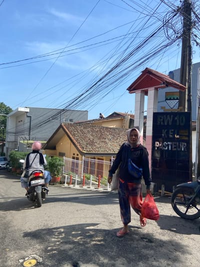 The image depicts a street scene in the Pasteur neighborhood of Bandung, Indonesia, located at coordinates -6.892 latitude and 107.599 longitude. A woman wearing a headscarf and carrying a red bag walks down the street towards a sign that reads "RW 10 KEL PASTEUR." A motorcycle with a pink helmet on the seat is parked to the left of the sign. The sky is clear blue with a few scattered clouds. There are numerous power lines extending from a utility pole in the background. There are two buildings in the background with tiled roofs. The street is paved with asphalt and has a slight incline. The scene is a typical street in a residential neighborhood.