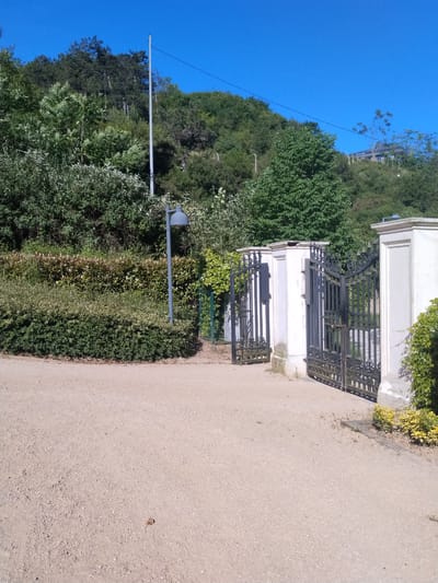 This image depicts an outdoor scene, likely an entrance to a property, situated at the base of a densely wooded hillside under a clear blue sky, in Sarıyer, Turkey.
The foreground is dominated by a light brown gravel path or driveway, covering approximately the lower half of the frame. A single dark, dried leaf or small organic debris is visible on the gravel towards the bottom center.
In the midground, a set of ornate, black wrought-iron gates, flanked by tall, white, rectangular pillars, are partially open, leading into a treed area. The gates feature intricate scrollwork and spiked finials. A grey lamppost with a downward-facing, bell-shaped lamp head stands to the left of the gates, positioned at the edge of the gravel path amidst green shrubbery. Dense, well-maintained green hedges and various types of deciduous and coniferous trees form a barrier and extend behind the gates. The white pillars show minor staining and wear at their bases. On the right pillar, green and yellow variegated foliage climbs slightly.
The background consists of a steep, heavily vegetated hillside covered with numerous trees, both broadleaf and coniferous. A slender grey pole, possibly a utility pole or antenna, rises prominently from the left side of the hillside. Overhead power lines stretch horizontally across the upper mid-section of the image, appearing to connect to structures on the hillside. On the far right of the hillside, a building with a dark roof and light-colored walls is partially visible, nestled among the trees. The sky above is uniformly clear and bright blue, indicating sunny conditions.
No people or animals are visible within the frame. There are no explicit actions or interactions depicted.