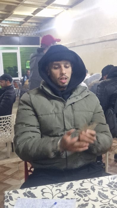 This image depicts an indoor scene in Fez, Morocco, featuring a male subject prominently in the foreground. The male has light skin, short dark curly hair partially visible beneath a black hoodie, and a light mustache and goatee. He is wearing an olive-green puffer jacket over a black hooded sweatshirt. His hands are positioned near his chest, appearing to manipulate a small, indistinct brown object. His mouth is slightly open.
The subject is seated at a table covered by a cream-colored tablecloth with a repeating dark floral or leaf pattern. On the tablecloth, to the subject's left, lies a handwritten ledger or score sheet containing multiple entries, along with a blue ballpoint pen.
In the background, several other individuals are present within the same establishment. To the left of the main subject, a man in a black jacket and black cap is seated, facing away from the camera. Further back, a person wearing a red baseball cap is partially visible. Another individual in a dark jacket is partially visible to the right. The interior environment includes light-colored walls, a ceiling with a grid pattern and integrated light panels, and a white-framed door or window in the distant left background. White plastic chairs with decorative cut-out patterns and patterned tiled flooring are visible, suggesting a casual public gathering space.