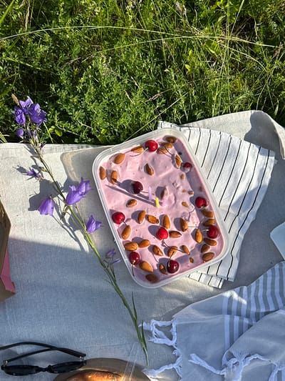 A clear plastic container filled with a pink, creamy substance, possibly ice cream or yogurt, is placed on a grey and white striped napkin. The container is garnished with whole almonds and cherries, some of which have stems. Two small, unlit birthday candles, one yellow and one pink, are stuck into the mixture. The container and napkin are situated on a light grey picnic blanket. To the left of the container, a stem of purple bellflowers with green leaves casts a shadow on the blanket. In the background, lush green grass and other vegetation are visible, suggesting an outdoor setting. In the foreground, a pair of black-rimmed sunglasses rests on the blanket. A brown paper bag is partially visible to the far left, and a white towel with fringed edges lies to the right of the container. The scene is illuminated by sunlight, creating distinct shadows. The location is the city of Olkhovka, Russia.