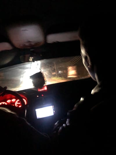 The image depicts the interior of a car at night. A person's head and shoulder are visible on the right side, silhouetted against the darkness. In the foreground, on the left, a glowing red dashboard with illuminated gauges is visible, indicating the car is operational. A car stereo or display screen is also visible in the center, emitting a bright white light. Outside the windshield, the scene is illuminated by streetlights and the headlights of the vehicle, revealing a road and some structures. A hanging air freshener is visible from the rearview mirror. The overall impression is of a night drive through a city, possibly Rukuba Road, Nigeria.