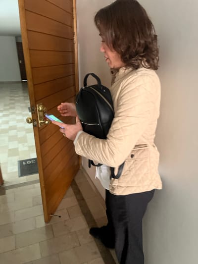 An adult female is standing in an indoor residential hallway, positioned next to a partially open wooden door. She has brown hair and is wearing a quilted beige jacket over a white long-sleeved shirt, black trousers, and black shoes. Her left hand holds a black backpack, from which a white tag or paper is visible. Her right hand is holding an illuminated smartphone, which she is orienting towards a brass-colored smart lock assembly on the door. The door, constructed with horizontal wood panels, is open inward, revealing a light-colored tiled floor with a "301" doormat immediately inside the threshold. The tiled floor continues into the background, showing an empty interior space. The precise location within Bogota, Capital District, Colombia, cannot be determined from the visual information presented.