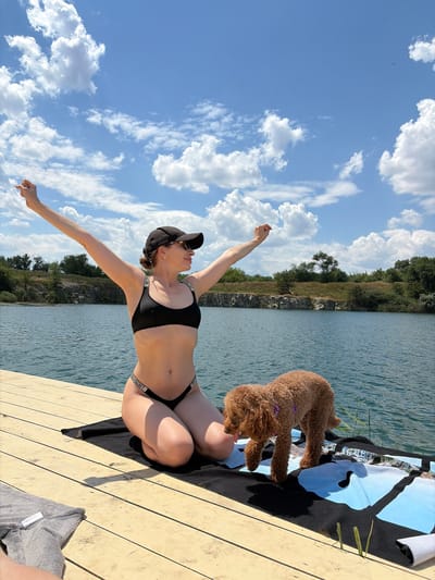 In Novomykolaivka, Ukraine, a woman in a black bikini and baseball cap sits on a wooden dock, her arms outstretched towards a bright blue sky filled with fluffy white clouds. Beside her, a small, curly-haired brown dog, wearing a purple collar, looks down at her, its tongue slightly out. The lake's calm surface reflects the surrounding greenery and the sky. The scene suggests a warm, sunny day, likely in the afternoon, given the strong shadows cast on the dock. The woman's posture conveys a sense of freedom and enjoyment. The bikini straps feature the text "VICTORIA'S SECRET" in repeating letters.