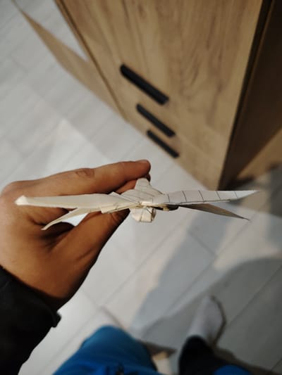 A person holds a white paper airplane in their hand, likely indoors. The background includes part of a wooden cabinet with black handles, a tiled floor, and a glimpse of the person's clothing - a blue shirt and a black sleeve. There is no information in the image to confirm that it was taken in Panagyurishte, Bulgaria. The image does not include any additional context.