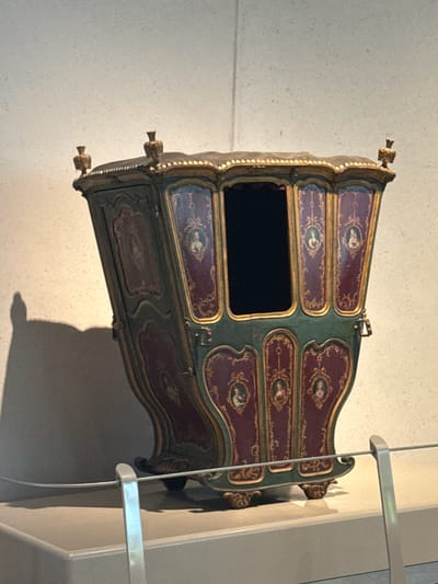 The image shows a decorative, antique sedan chair displayed against a wall. The chair is predominantly maroon and green with gold trim and features painted portraits within decorative frames. It has a dark, rectangular opening where the occupant would sit. The sedan chair is positioned near a metal railing or barrier. The location is not evident from the image, so San Bernardino Tlaxcalancingo, Mexico, cannot be confirmed.