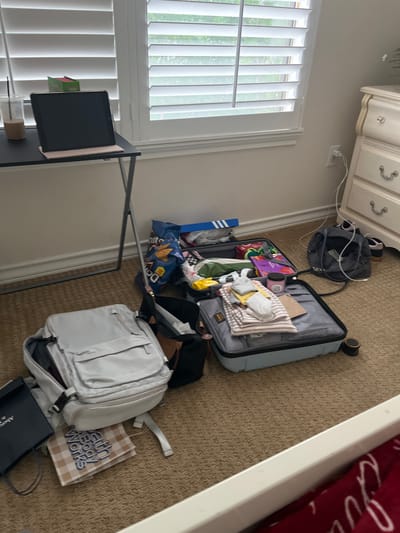 The scene is indoors, likely a bedroom, cluttered with luggage and travel-related items. An open suitcase lies on the carpeted floor, filled with clothes and various personal belongings. Next to it, there is a white backpack and shopping bags. A black backpack is also present. A desk with a tablet is positioned near a window with blinds. A dresser is partially visible in the upper left corner of the frame. The location appears to be a private residence. There is no evidence to suggest it is in San Antonio, United States of America.