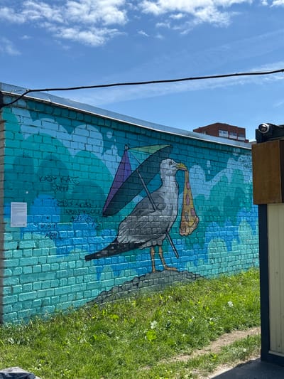 This image captures a vibrant mural painted on a brick wall, depicting a stylized heron or stork. The bird is shown with its beak holding a slice of pizza, and it is also carrying a folded umbrella with purple and black panels. The background of the mural is a gradient of blues and greens, suggesting water or a sky, with circular patterns that could represent bubbles or ripples.

Graffiti is visible on the mural, with several lines of text in black spray paint. One prominent tag reads "KADJA KAS OIMAD". Another tag, partially obscured, appears to be "BUSZOKFU". There is also a small, framed information plaque at the bottom center of the mural, which is illegible in this resolution.

Above the mural, a bright blue sky with scattered white clouds can be seen. A black electrical cable runs diagonally across the sky and attaches to the wall near the top. A security camera is mounted on a wooden overhang at the top of the image, and a tall brick building with visible windows is present in the background to the left, behind the mural. To the right of the mural, a patch of lush green grass with some small white flowers growing in it is visible.

The lighting suggests it is daytime, likely sunny given the bright sky and shadows. The overall impression is a whimsical and somewhat surreal street art piece displayed in an urban outdoor setting.