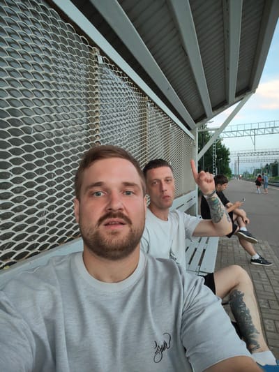 A selfie taken at a train station platform in Luga, Russia, shows two men sitting on a bench. The man in the foreground, closer to the camera, is wearing a grey sweatshirt and has a beard. The man behind him, to his right, is wearing a white t-shirt and pointing his index finger upwards. He has a tattoo on his left arm. In the background, other people are visible on the platform, and train tracks and overhead wires indicate the setting. The station has a metal mesh fence and a covered roof supported by white pillars. The sky is overcast.