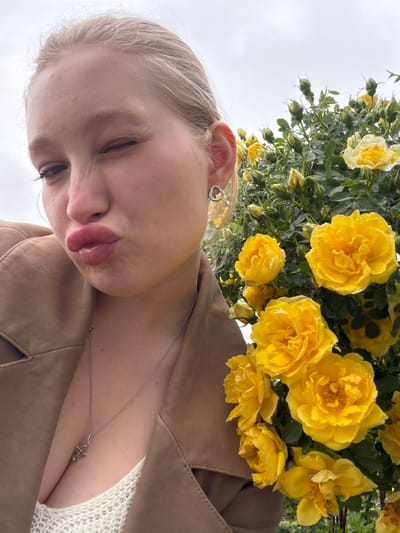 A close-up, slightly angled photograph shows a person with light hair and skin, positioned against a backdrop of yellow flowers. The person is wearing a brown jacket over a white, textured top. A silver necklace with a pendant is visible. The flowers are predominantly yellow roses, some with water droplets on the petals. The background appears to be an overcast sky, indicating outdoor setting. The location is not discernible from the image alone.