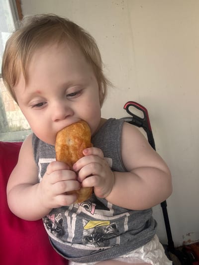 A young child with blonde hair and a red mark on their forehead is sitting and eating a piece of food, likely bread or pastry, with both hands. The child is wearing a grey tank top with a graphic print of cats and yellow stripes, and a white diaper. They are seated on a red surface. In the background, a light-colored wall is visible, with a black and red vacuum cleaner handle leaning against it. The scene appears to be indoors. This image was captured in the city of Sayanogorsk, Russia.