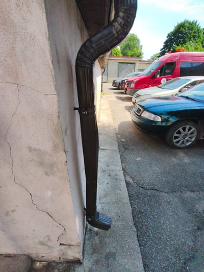 A downward-facing shot shows a black downspout attached to a cracked, pale stucco wall in Tukums, Latvia. The downspout curves sharply and extends out of frame at the bottom. In the background, parked cars and a red van are visible against a backdrop of trees and a bright sky. The scene appears to be an outdoor urban environment.