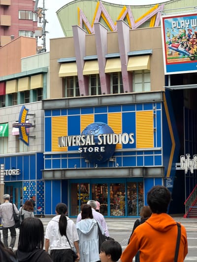 This media file captures a bustling street scene in Osaka, Japan, featuring the vibrant facade of a Universal Studios store. The central building is characterized by bold blue and yellow paneling, with a prominent sign reading "UNIVERSAL STUDIOS STORE" in silver lettering, accompanied by a large blue globe graphic. Below this, another section of the building also features blue walls adorned with colorful star and dot patterns, and displays the word "STORE."

To the left, an adjacent building has a more neutral beige and light brown palette, with distinct angled window awnings. High above this structure, a large poster is visible, advertising "PLAYWORLD" with colorful cartoon characters that resemble iconic video game figures. On the upper right, part of a sign that appears to say "GLYSS" or similar is partially visible.

A number of people are seen in the foreground and middle ground, mostly from behind, walking along a paved outdoor area. They are casually dressed and appear to be visitors or shoppers, moving past the storefronts. Some individuals seem to be looking into the shops, suggesting a retail or entertainment complex setting. The time of day appears to be daytime, with soft, diffused lighting. The overall scene conveys a lively, commercial atmosphere, consistent with a major tourist destination like Universal Studios Japan.