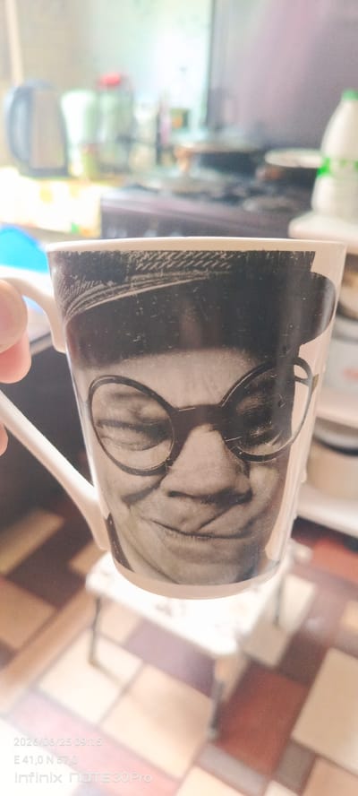 A close-up shot shows a white ceramic mug with a black and white image of a person wearing glasses and a hat. The person's eyes are closed, and they appear to be smiling. The mug is held by a person's hand, which is visible on the left side of the frame. The background is out of focus and appears to be a kitchen setting with appliances and cabinets. A timestamp and location information in the bottom left corner of the image indicates the photo was taken on June 25, 2026, at 09:15, at coordinates E 41.0 N 57.0, using an Infinix NOTE 30 Pro phone. This location appears to be in the city of Ivanovo, Russia.