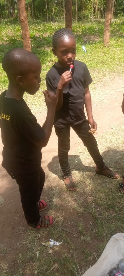 Two young boys stand in a grassy clearing in the forest. The boy on the left is wearing a black shirt with "PEACE" written on it and black shorts. He is wearing red sandals. The boy on the right is wearing a black shirt and jeans, and brown sandals. He is holding a red lollipop in his right hand. The boys are standing on a dirt path. The forest is in the background. There is a tree on the left side of the image. The location is a forest clearing in Lwandeti, Kenya, near the coordinates (latitude: 0.579, longitude: 34.808).