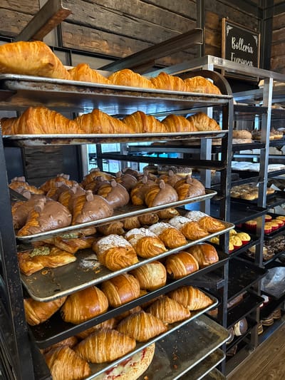 The image depicts a bakery display case filled with various types of croissants. The croissants are arranged in metal trays. Some are plain, others are covered in chocolate and topped with small chocolate pieces, while others are sprinkled with powdered sugar or filled with pistachio cream. The display case is made of black metal and has a sign that reads "Bolleria Europea". The location is a bakery in Puebla, Mexico.