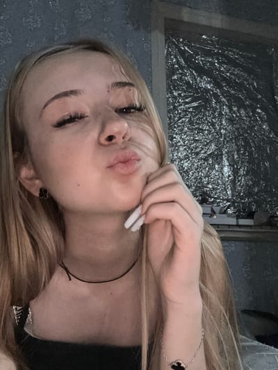 The image shows a young woman with blonde hair, taking a selfie. She is wearing a black top and a black choker necklace. She is also wearing a bracelet with a clover-shaped charm. Her right hand is raised to her face, and her left hand is not visible. She has her lips pursed as if to kiss the camera. The background appears to be a room with blue patterned wallpaper and a large reflective surface. The location is not specified within the image.