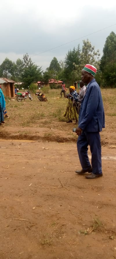 The scene takes place in a rural area of Lwandeti, Kenya, with coordinates 0.554 latitude and 34.806 longitude. It is a dirt road with a man in a blue suit and a red and green cap walking toward the camera. He is holding a black cellular device in his right hand. There is a woman in a yellow and green dress and a man in a white shirt and blue pants walking away from the camera. Two motorcycles are parked in the background. It is a cloudy day with scattered trees and a few buildings. The environment is dusty and dry. The scene appears to be a casual outdoor gathering in a rural area.