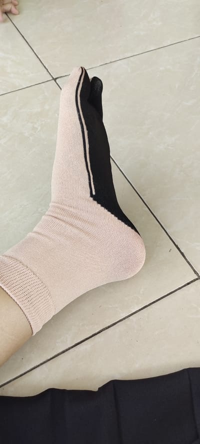 The image shows a person's foot wearing a light brown sock with a black stripe running down the side. The sock is partially pulled up, revealing a small portion of the person's leg. The person's leg is resting on a white tile floor. There is a black piece of fabric in the lower right corner of the image. The coordinates -7.443, 112.71 correspond to a location in Sidoarjo, Indonesia. The image does not show any identifiable faces or other people. The image suggests that someone is getting dressed or preparing for an activity.