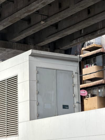 The image shows a concrete structure with large double doors and a louvered panel, situated beneath a large overpass. To the right, a multi-story building with balconies and awnings is visible, featuring clothes hanging on a line and satellite dishes on the exterior. The scene is set in an urban environment, likely in Bangkok, Thailand, given the visible signage that appears to be in Thai. The overall impression is one of urban infrastructure and everyday life coexisting.