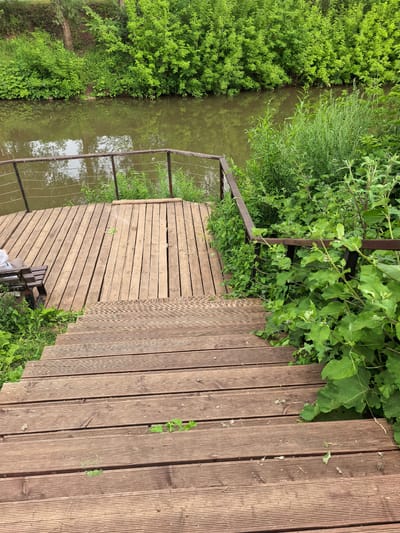 The image depicts an outdoor scene featuring a wooden deck and stairs leading down towards a body of water bordered by lush green vegetation. The deck, constructed from horizontal wooden planks, has a metal railing supported by vertical posts and thin wires. A wooden bench with a dark seat is visible on the left side of the deck. The water appears calm and reflects the surrounding greenery. The vegetation is dense and consists of various types of plants, including large-leafed plants and tall grasses. The overall environment suggests a natural or park-like setting. The specific location in the city of Sterlitamak, Russia, is not discernible from the image itself.