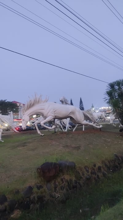 The image captures a public art installation featuring multiple white horse sculptures in motion, likely located in the city of Pusakarakyat, Indonesia. The sculptures are placed on a grassy mound, suggesting a park or a prominent roadside attraction. In the background, there are trees, buildings, and parked vehicles, indicating an urban or semi-urban setting. Several power lines are visible against the overcast sky, adding to the context of a developed area. The foreground shows a grassy slope with some rocks, and a ditch or drainage area. The lighting suggests it is either early morning or late evening. There are no people visible in the image.