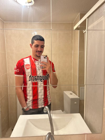 In a mirror selfie, a man stands in a bathroom with beige tile walls. He is wearing a red and white soccer jersey and jeans, holding his phone. The reflection shows a toilet, sink, and faucet. A medicine cabinet with a light above is visible. The man is smiling slightly. The scene is brightly lit.