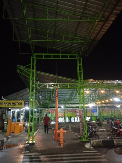 The media file captures an outdoor scene at night, likely a public or commercial area, characterized by extensive green metal structures and corrugated roofs providing shelter. The setting appears to be a parking or commercial complex in Kecamatan Kartasura, Indonesia.
In the midground, a man wearing a red shirt is walking away from the camera, traversing a paved area with white striped markings resembling a pedestrian crossing. A prominent orange pole, possibly a street light or utility pole, with a fire hydrant-like base, stands near him.
To the left, a yellow sign clearly displays "PARKIR MOTOR CUSTOMER," indicating a motorcycle parking facility. Below it are yellow barriers or gates, suggesting an entrance or exit point, with the word "KELUAR" (meaning "exit" in Indonesian) also visible. The area around the parking entrance is artificially lit.
To the right, beyond the central green framework, several motorcycles are parked, indicating active use of the parking area. More green metal roofing and support structures extend into the background, where string lights and other illuminated features suggest a food court or market area. Partially visible text reads "FOOD GAI," likely part of a business name or sign.
The time of day is night, with the sky completely dark. Artificial lighting, including overhead lights under the roofs and more decorative string lights in the distance, illuminates the scene. There are no clear indications of weather conditions, but the ground appears wet in places, possibly from recent rain or reflections. The overall atmosphere is functional and active, typical of a bustling commercial or public space after dark.