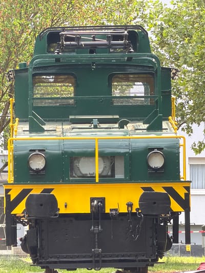 The image shows a close-up, head-on view of a green and yellow diesel locomotive. The front end is primarily green with yellow accents, including stripes and handrails. The locomotive's front has black diagonal stripes against a yellow background. Headlights are visible. Behind the locomotive, a building with windows can be seen, and foliage, including trees and grass, is present. The scene is likely located in Esch-sur-Alzette, Luxembourg.