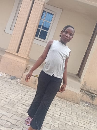 A young female child stands outdoors in front of a building in Zango-ndaji, Nigeria. The child wears a light grey sleeveless top, black trousers, and light purple sandals. Her dark hair is styled in braids or cornrows. She is positioned slightly off-center, facing the camera with a neutral expression. Behind her is a light beige building facade featuring a fluted architectural column on the left and a white-framed window with blue-tinted, gridded panes. The ground is paved with light grey interlocking concrete blocks. A dark brown architectural element is visible on the far right. The scene is illuminated by natural daylight.