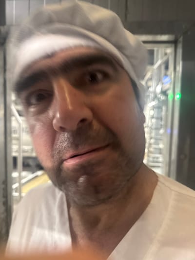 The image is a close-up, blurry photograph of a person wearing a white chef's hat and white shirt. The person appears to be male with dark hair and a beard. He is looking directly at the camera with a surprised or concerned expression. The background is out of focus but appears to be an industrial or commercial kitchen setting with metal shelving and some lights. There is no specific information within the image that would indicate the location in Dedovsk, Russia.