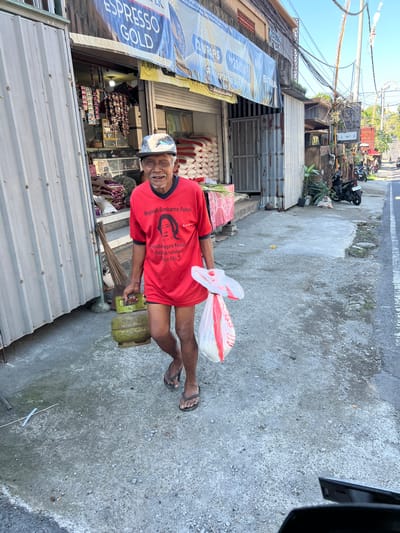 An elderly Indonesian man, wearing a red t-shirt with a portrait of Megawati Soekarnoputri and a patterned baseball cap, walks down a sidewalk in Sukawati, Indonesia. He is carrying a green propane tank in his left hand and a white plastic bag filled with white goods in his right hand. The man is barefoot, wearing dark-colored flip-flops. The sidewalk is made of concrete and is lined with shops. One shop has stacks of rice bags and a display of goods. The street in front of the shops is paved, and several motorcycles are parked along the side of the road. The sky is clear and blue, indicating a sunny day.