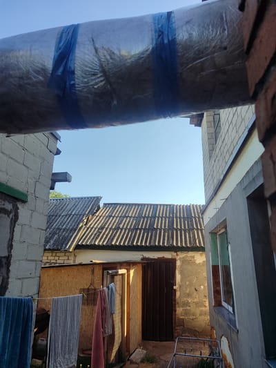 The image captures an outdoor scene, likely a backyard or alleyway, under a clear blue sky. In the foreground, a large, insulated pipe runs horizontally across the top of the frame, secured by blue tape. To the left and right, unfinished or under-construction brick buildings frame the view. The left building is made of white cinder blocks, with a section of green trim visible. The right building is a lighter gray, also constructed with cinder blocks, and features a window with a dark frame.
In the midground, several corrugated metal roofs of what appear to be outbuildings or smaller structures are visible. One of these structures has a brown metal door. A clothesline is strung between two points, with several towels and clothing items hanging to dry, including a blue towel on the left, a gray towel in the center, and a pink towel to its right. A small metal drying rack with clothespins is also present on the ground near the right side. The ground appears to be a mixture of dirt and possibly some patchy grass. The overall impression is one of a functional, possibly residential, area undergoing some level of maintenance or construction. The scene does not explicitly indicate a specific location within Cherkasy, Ukraine, beyond being an outdoor setting. There are no people visible in the image.