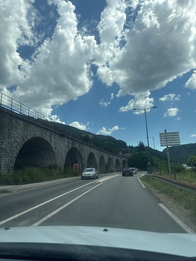 This is a vertical, eye-level, medium shot taken from a vehicle. The scene depicts a road with a viaduct on the left and lush green hills in the background. The sky is bright blue with dramatic white clouds, suggesting a sunny day.
The viaduct is constructed of large, grey stones and features a series of arched openings. A metal fence runs along the top of the viaduct. To the right of the road, there is a green, grassy embankment with some trees. Several cars are visible on the road, suggesting traffic. A tall lamppost with two lights stands near the road.
A prominent signpost on the right side of the road displays several directional arrows and French text, including "Sur Le Puits-Villedieu," "Gare S.N.C.F & Routière," "Gendarmerie PGM," "Crèche Municipale," "La Maison," and "France services." This text provides location context, indicating the presence of services and destinations within the Hauts de Bienne region of France.
The overall impression is a scenic drive through a mountainous or hilly area on a pleasant day. The image captures a moment of travel, with a focus on the infrastructure and natural beauty of the location. The perspective from inside the vehicle adds a sense of movement and personal experience to the scene.
