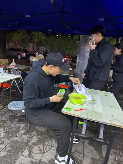 The image shows a group of people in what appears to be an outdoor setting in Zhongtun, China. Two men are standing at a table where food items like a green bowl and plastic bags are visible. One of the men is looking at his mobile phone. A third man is sitting in a chair at another table and appears to be eating or unwrapping food. A blue tent structure is on the left side of the frame. A car is visible in the background. The ground is paved with stone or concrete.