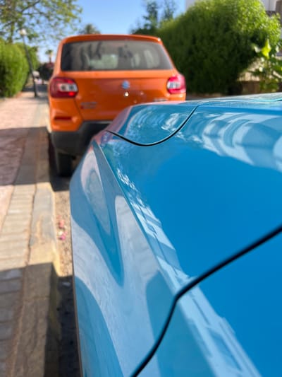 The image shows a close-up, low-angle view of the hood of a bright blue car, with the rear of an orange car visible in the background. The blue car's hood reflects the sky and surrounding environment, showing curves and a seam line. The orange car is parked on a paved surface next to a sidewalk, with greenery and trees in the background. The lighting suggests it is daytime, with sunlight casting shadows. The scene is consistent with a residential or public parking area in Sharm El Sheikh, Egypt, a resort city known for its sunny climate and coastal environment. No people are visible in the image.