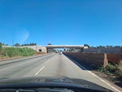 The image shows a view of a paved highway in Nduma, Kenya (latitude: -1.181, longitude: 36.644). The highway has a concrete overpass running perpendicular to the road. The overpass is in the background, partially obscured by foliage. The image appears to have been captured from a vehicle traveling on the highway, as a black car's windshield and dashboard are in the foreground. The sky is clear blue with minimal clouds. There is a small amount of greenery and trees along the sides of the road, with more trees in the distance. Two vehicles can be seen in the distance driving on the highway. The vehicles are a white car and a truck. The road is empty in the foreground. The highway is a four-lane road. The left shoulder is unpaved. The right shoulder is paved with a concrete barrier. The road is relatively straight, with no visible curves or turns. The image appears to have been captured during the day, as there is no indication of nighttime lighting.