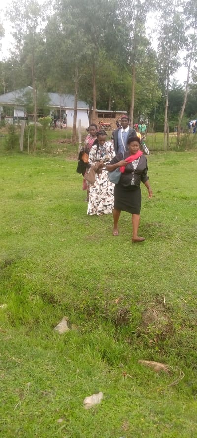 The image shows a group of five people walking on a grassy field. They are all adults and are dressed in casual clothing. The person in the front is wearing a black skirt and a red scarf, and the other four are wearing dresses or shirts. There are trees and bushes in the background, and a building can be seen in the distance. The location of the image is Kivaywa, Kenya, which is situated at the coordinates 0.56 latitude, 34.806 longitude. The sky is overcast. There are some branches and leaves on the ground near the bottom of the image.