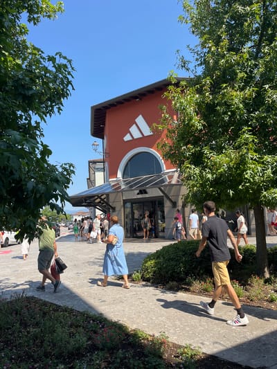 This image depicts the exterior of an Adidas outlet store in the city of Rodengo, Italy, under a clear blue sky on a sunny day. The store is a modern, reddish-orange building with a large, arched window entrance above a glass awning. Prominently displayed on the facade is a large white Adidas logo. Several people are walking on a paved walkway in front of the store, some carrying shopping bags. Trees with green foliage frame the scene on both the left and right sides, casting shadows on the ground. In the background, other buildings and cars are visible, suggesting an outdoor shopping complex. The people are dressed in casual summer attire. A man in a black t-shirt and yellow shorts is walking towards the viewer's right, and a woman in a blue dress is walking towards the viewer's left. The ground is a light-colored, speckled pavement with a border of green bushes and small red and pink flowers.