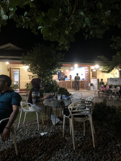 This is a nighttime photo taken outdoors in Tambonbaroh, Indonesia, likely at a cafe or restaurant. The scene is dimly lit by artificial lights, with some areas well-illuminated and others in shadow. Lush green foliage frames the left side of the image, contrasting with the dark night sky.
The main focus is on a seating area with several white plastic chairs and a few tables. In the foreground, a person wearing a dark green shirt is partially visible, with their arm extended towards a chair. Further back, a person wearing a headscarf and a striped shirt is seated at a table, looking towards their laptop. Behind them, there are more chairs and tables, some of which appear occupied by other individuals.
Through an open doorway in the background, a brightly lit interior can be seen, where several people are standing and working behind a counter made of wood. This suggests the establishment is a cafe or food service area. The ground is covered with small, dark grey pebbles, characteristic of an outdoor patio or garden setting. The overall mood is relaxed and casual, with the warm interior lighting creating an inviting atmosphere. There is no visible text in the image.