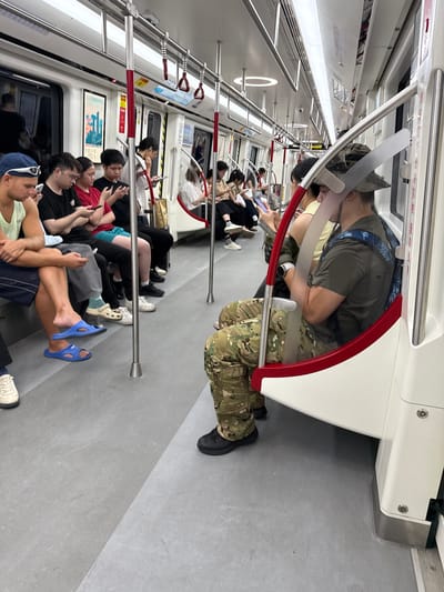The image shows the interior of a subway train in Baiyun District, China, with several passengers seated and interacting with their mobile phones. In the foreground on the left, a man with a blue cap and sunglasses is looking at his phone. Beside him, a group of people are seated, also engrossed in their phones. On the right side of the train, a man wearing a camouflage hat and pants is visible, looking down at his phone. The train carriage is modern, with overhead handrails and posters on the walls. The lighting is bright, typical of public transportation. The scene depicts a common urban experience of individuals engaged with technology while traveling.