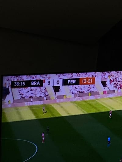This image depicts a digital screen, likely a television, displaying a soccer match. The scene originates from an interior location in the city of Meknes, Morocco. A scoreboard banner at the top of the screen shows the elapsed time as "36:15." The team identified as "BRA" has scored "3" goals, while the team "FER" has scored "0" goals, with an additional score of "(3-2)" listed next to "FER." The lower portion of the screen shows a green soccer pitch with white field markings. At least four players are visible on the field, one in a light uniform with a shadow, another in a red uniform, one in a dark uniform, and a fourth in a blue uniform. Spectators in stadium stands, appearing as blurred white and red masses, are visible behind the field. The ambient environment surrounding the screen is dark, emphasizing the illuminated display.